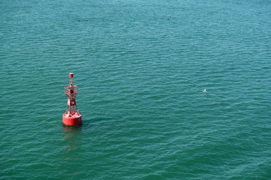 A red navigational buoy floating in calm blue ocean water under clear skies.